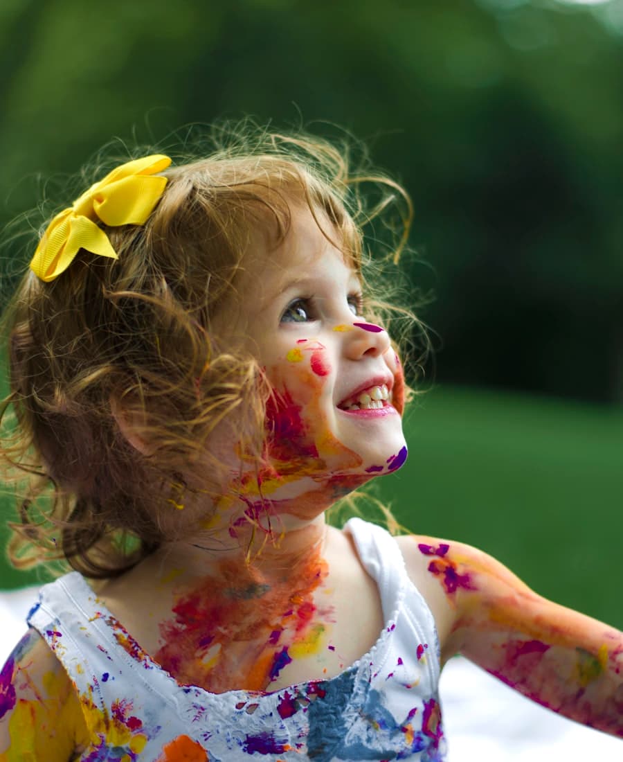 A young child smiling, her hands and face dotted with paint.